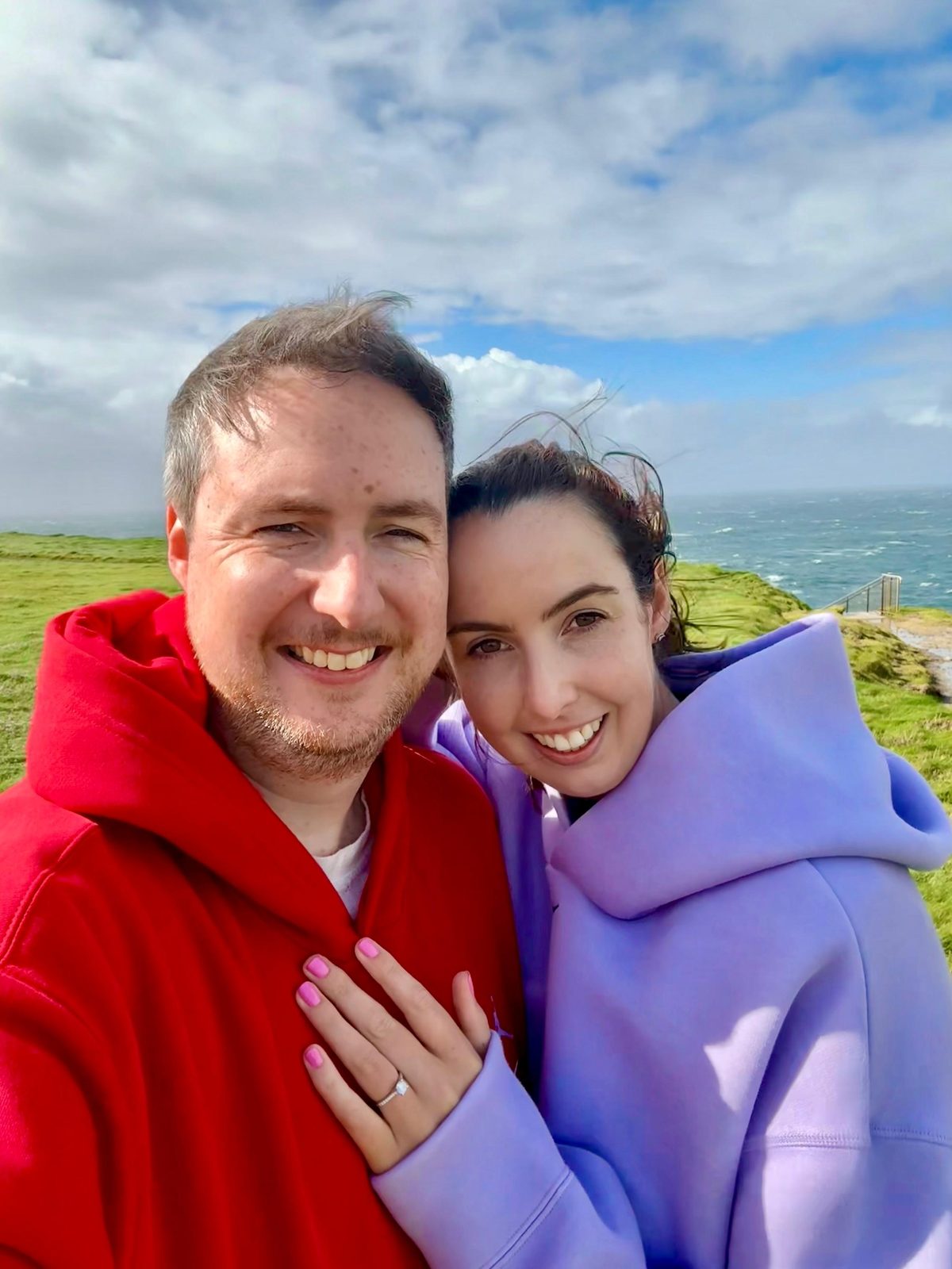 Orlaith and Niall cheek to cheek on the clifftop at Kilkee minutes after the proposal, her hand on his chest showing the ring, the Atlantic and a wild sky behind.
